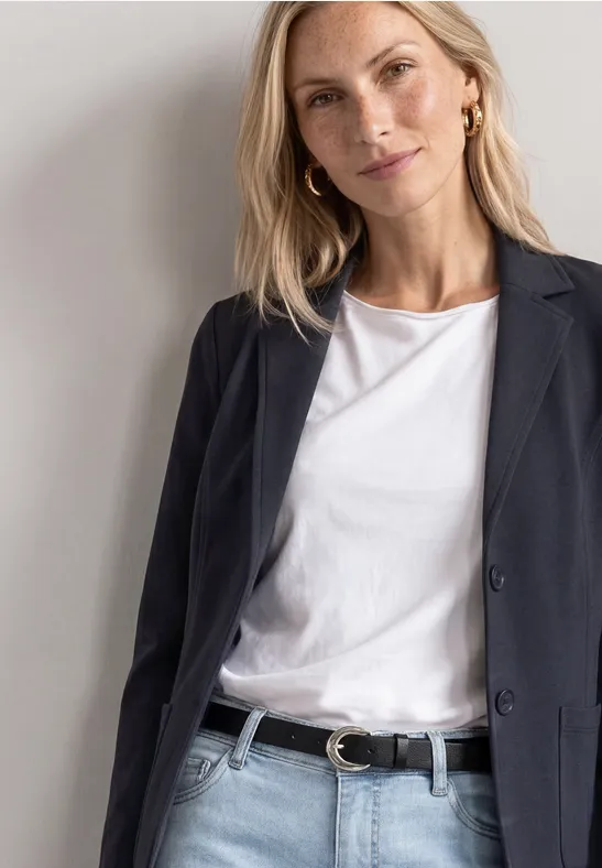 Eine Frau trägt einen schwarzen Blazer, ein weißes T-Shirt und einen schwarzen Gürtel vor hellem Hintergrund.