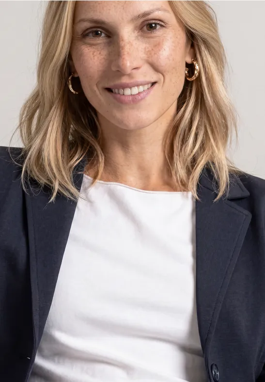 Lächelnde Blondine mit weißen T-Shirt und dunklem Blazer vor neutralem Hintergrund.