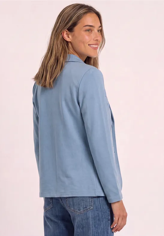 Een vrouw in een blauwe blazer en spijkerbroek kijkt glimlachend over haar schouder