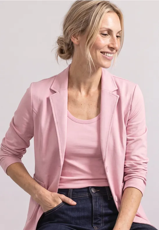 Lächelnde Frau in rosa Blazer und Top, mit Jeans und Hand in der Tasche.