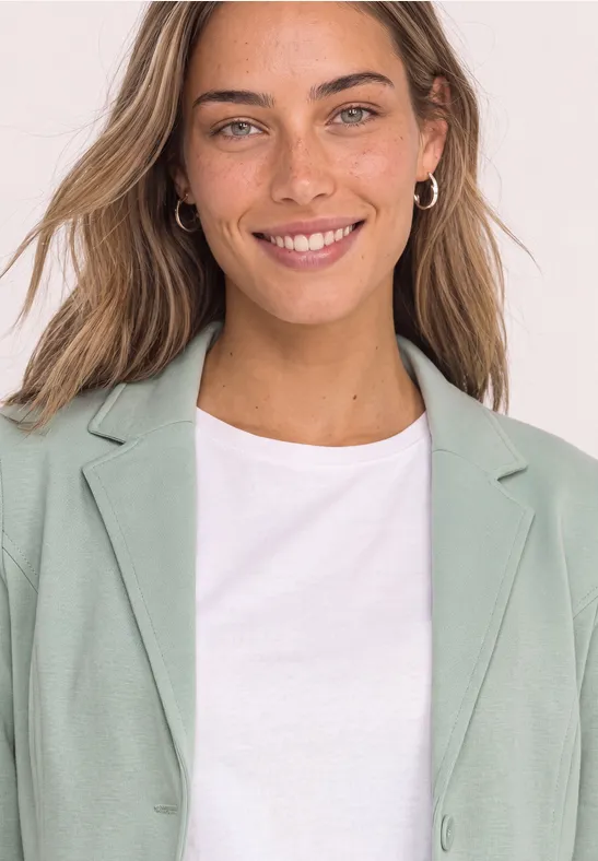 Eine lächelnde Frau in weißem T-Shirt und mintgrünem Blazer, mit schlichten Ohrringen vor hellem Hintergrund.