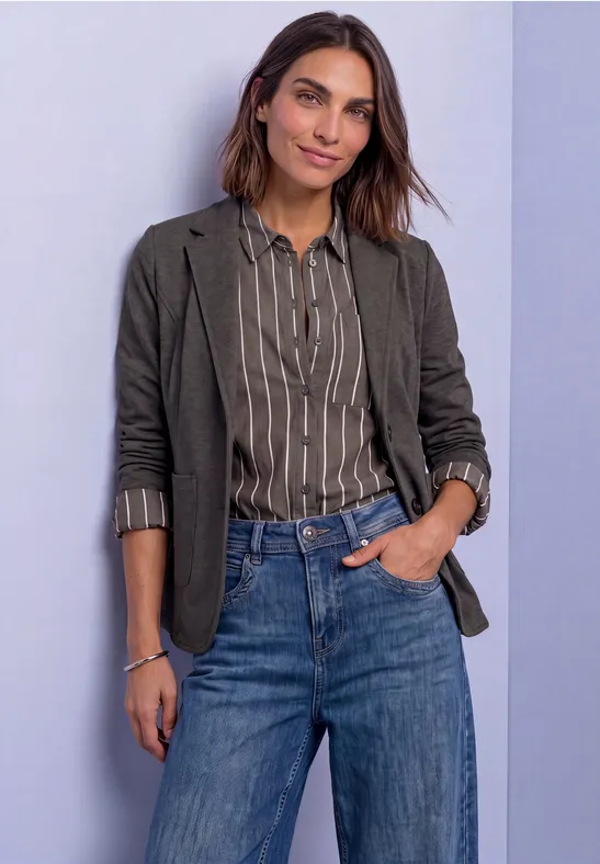 Frau in gestreifter Bluse, grüner Jacke und Jeans vor hellem Hintergrund.