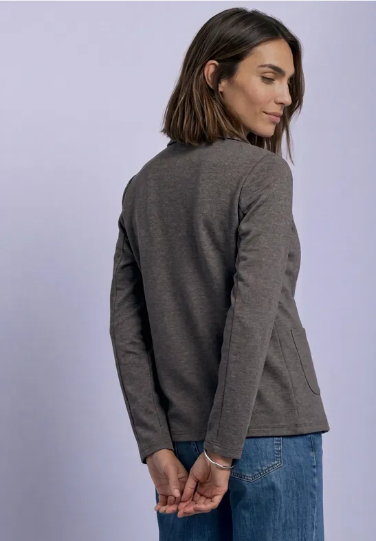 Junge Frau von hinten in grauem Langarmshirt und Jeans, Hände hinter dem Rücken verschränkt.
