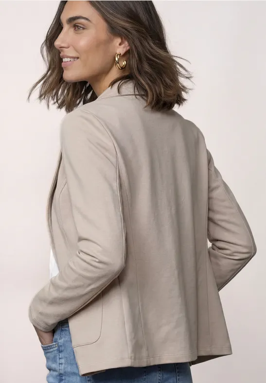 Frau in beige Blazer schaut über die Schulter, Hände in den Taschen, leicht lächelnd.