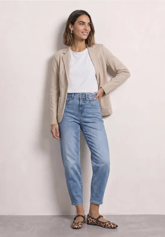 Frau trägt beige Blazer, weißes T-Shirt, blaue Jeans und Leoparden-Sandalen vor hellem Hintergrund.