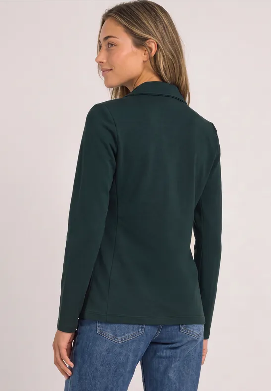 Frau trägt dunkelgrünen Blazer und Jeans, steht vor hellem Hintergrund, Blick über linke Schulter.