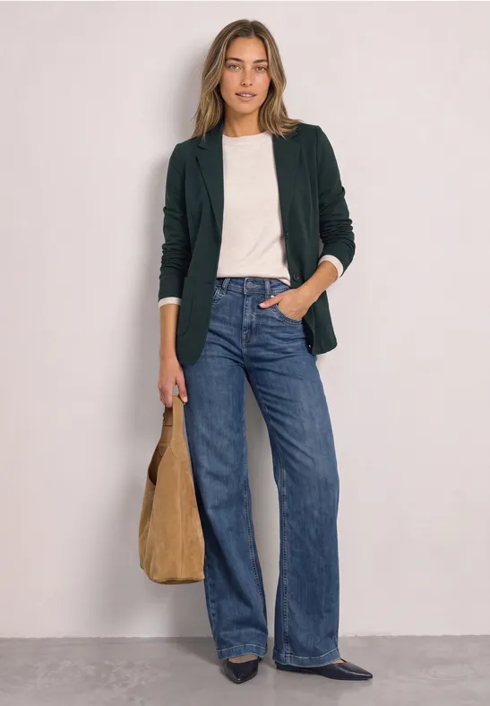 Frau trägt weißen Pullover, dunkelgrünen Blazer, blaue Jeans und hält braune Tasche