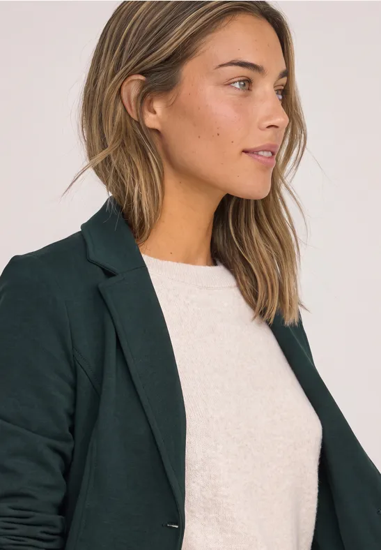 Frau mit kurzem hellbraunem Haar in dunkelgrünem Blazer und weißem Shirt, seitlich vor hellem Hintergrund.