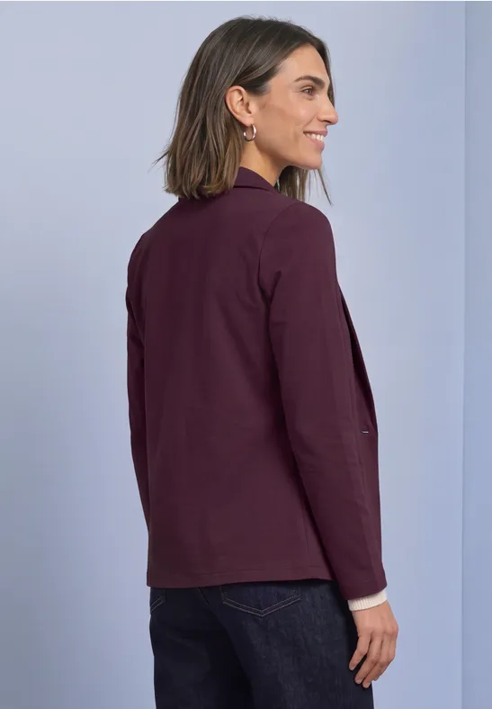 Eine Frau im Profil trägt einen burgunderfarbenen Blazer über dunkler Jeans vor hellem Hintergrund.