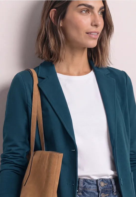 Frau trägt grünen Blazer, weißes Shirt und braune Ledertasche, Blick in die Ferne.