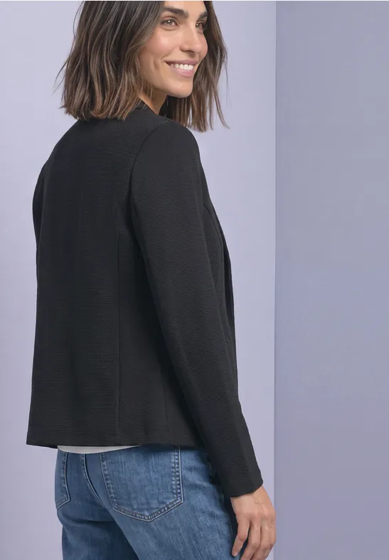Frau im schwarzen Blazer, schaut lächelnd über die Schulter, in Jeans vor pastellfarbener Wand.