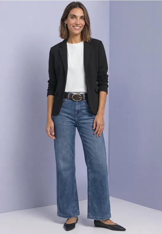Frau in schwarzem Blazer, weißem Shirt, weitem Jeans und schwarzen Ballerinas vor lila Hintergrund.