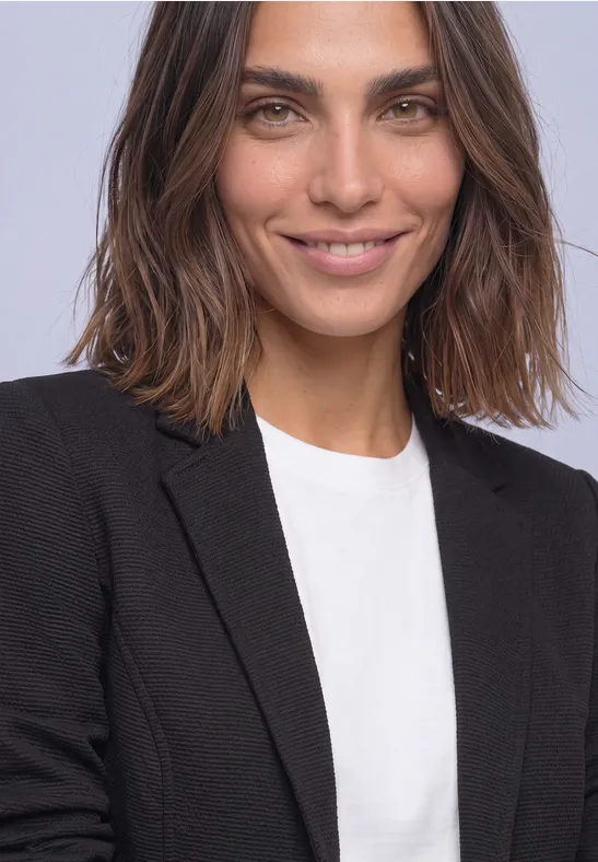 Eine lächelnde Frau mit schulterlangen braunen Haaren, die ein weißes T-Shirt und schwarzen Blazer trägt.