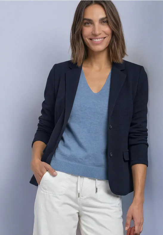 Frau trägt marineblauen Blazer, blauen V-Pullover und weiße Hose, lächelt vor grauem Hintergrund.