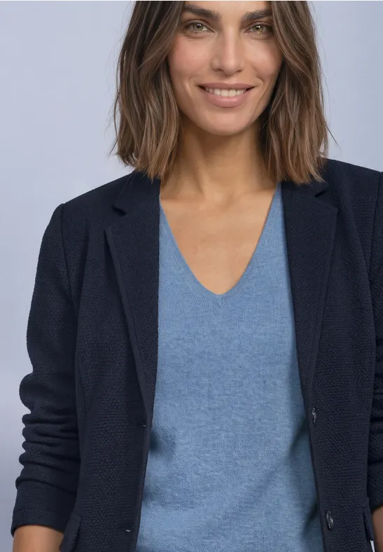 Frau mit brauen, schulterlangen Haaren, blauem V-Ausschnitt-Pullover und dunkelblauem Blazer, leichtes Lächeln.