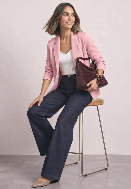 Frau in rosa Blazer, weißem Top und weiten Jeans sitzt auf Hocker und hält bordeauxrote Tasche.