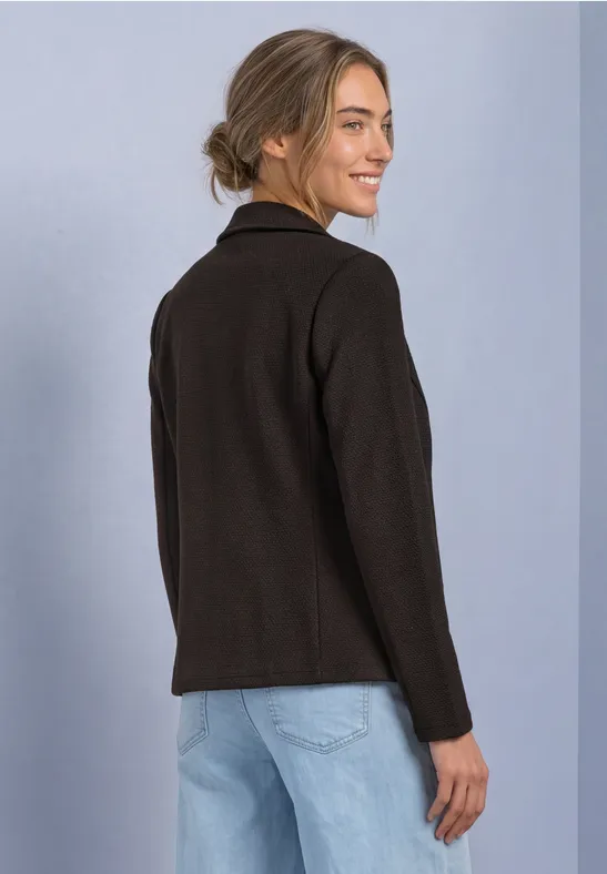 Femme en veste noire et jean, de profil, regarde par-dessus l’épaule et sourit.