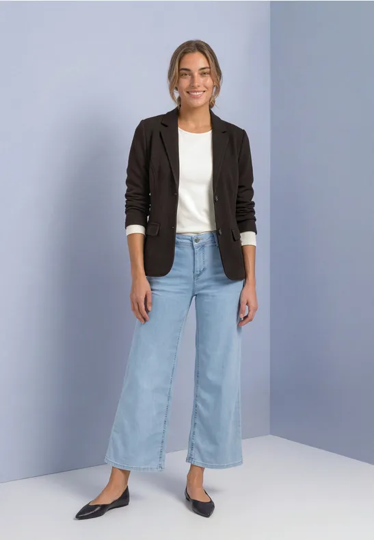 Jeune femme en blazer noir, t-shirt blanc, jean large et chaussures noires, posant devant un mur clair.