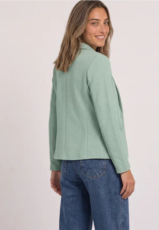 Frau trägt hellgrünen Blazer und blaue Jeans, steht mit Blick über die Schulter, lächelt vor neutralem Hintergrund.