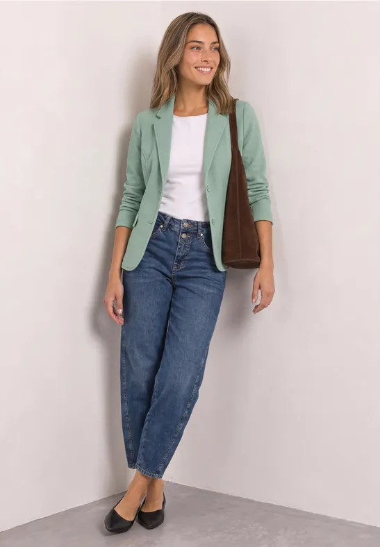Frau in grünem Blazer, weißem Top, Jeans und schwarzen Ballerinas, mit brauner Schultertasche vor hellem Hintergrund