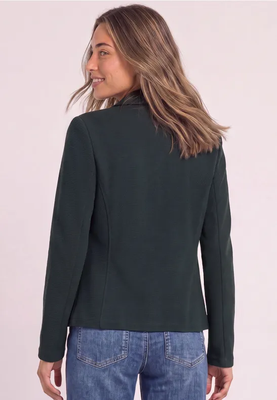Eine Frau von hinten in einem schwarzen Blazer und blauen Jeans vor neutralem Hintergrund.