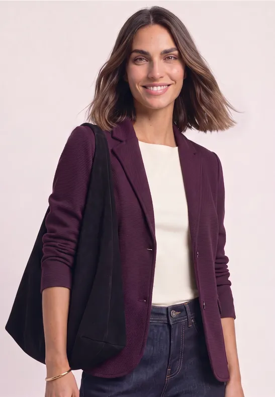 Junge Frau lächelt, trägt lila Blazer, weißes Shirt, Jeans und schwarze Tasche.