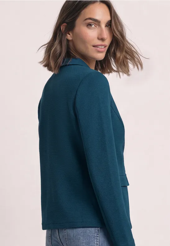 Frau in einem teal-blauen Blazer blickt seitlich über die Schulter.