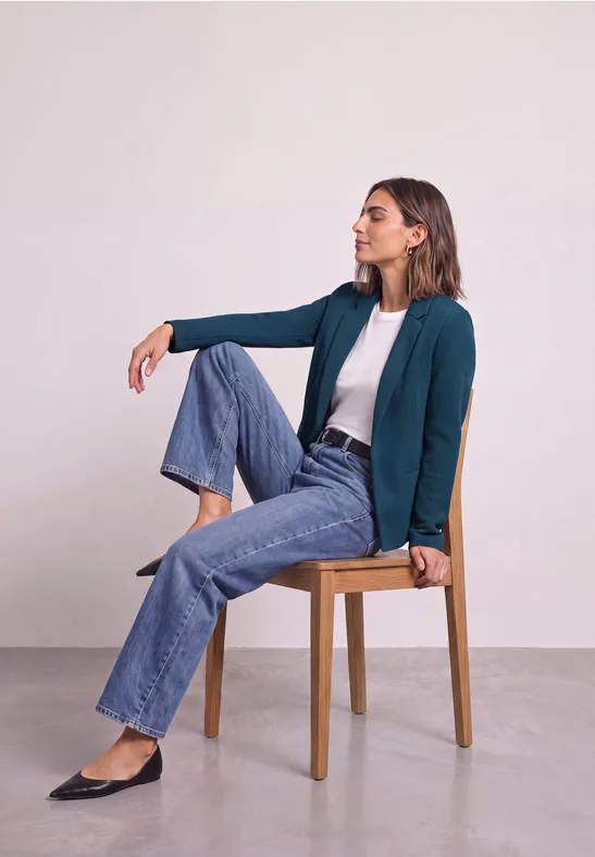 Frau sitzt lässig auf einem Holzstuhl, trägt Jeans, weiße Bluse und grünen Blazer.