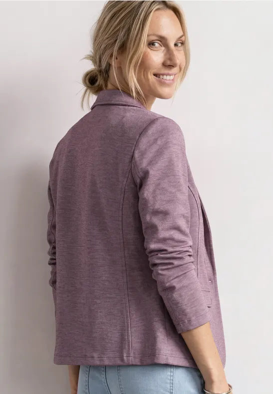 Person im lila Blazer, seitlich stehend, Blick nach links, Haar locker zusammengebunden.