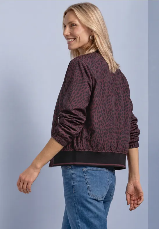 Blondynka w wzorzystej kurtce bomber i dżinsach uśmiecha się, spoglądając przez ramię.