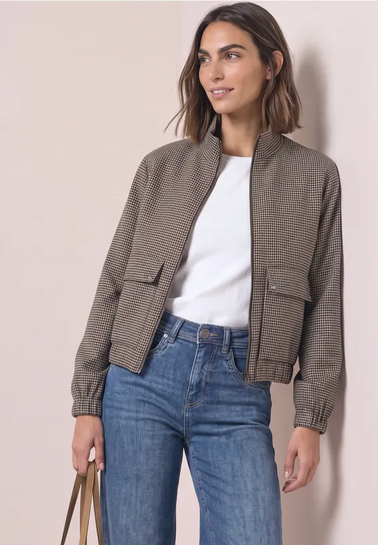Femme portant une veste bomber taupe, t-shirt blanc et jean, sac à main sur fond clair.