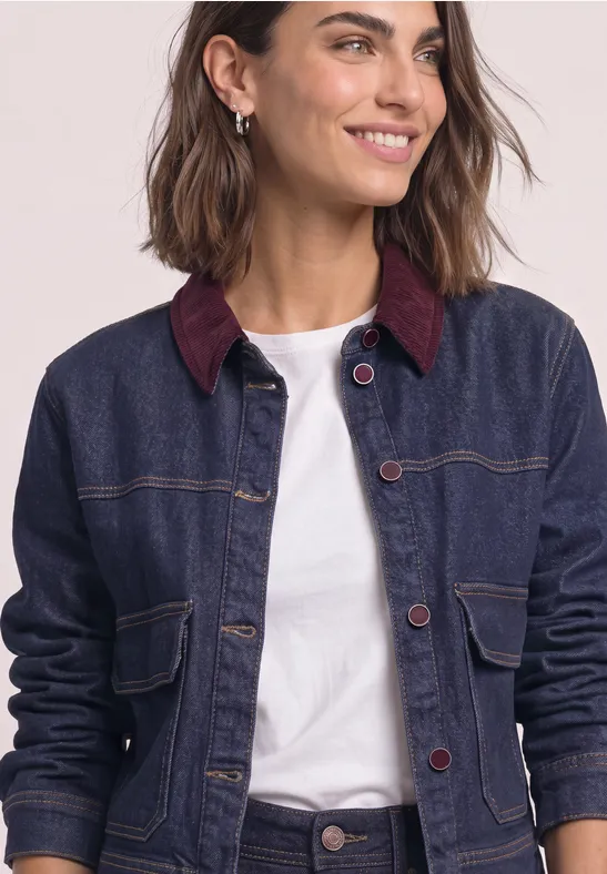 Femme souriante portant une veste en jean bleu foncé à col bordeaux et un T-shirt blanc sur fond clair.