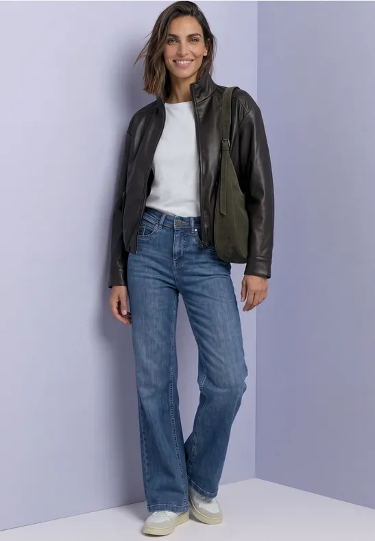Frau in Jeans, weißem T-Shirt, zweifarbiger Lederjacke vor lila Hintergrund.