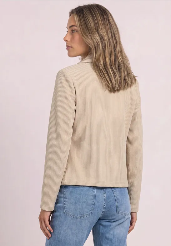 Frau steht im Profil, trägt eine hellbeige Jacke und blaue Jeans.