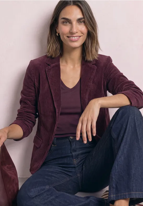Frau sitzt lächelnd, trägt ein weinrotes Sakko, dunkles Top und weite Jeans vor hellem Hintergrund.