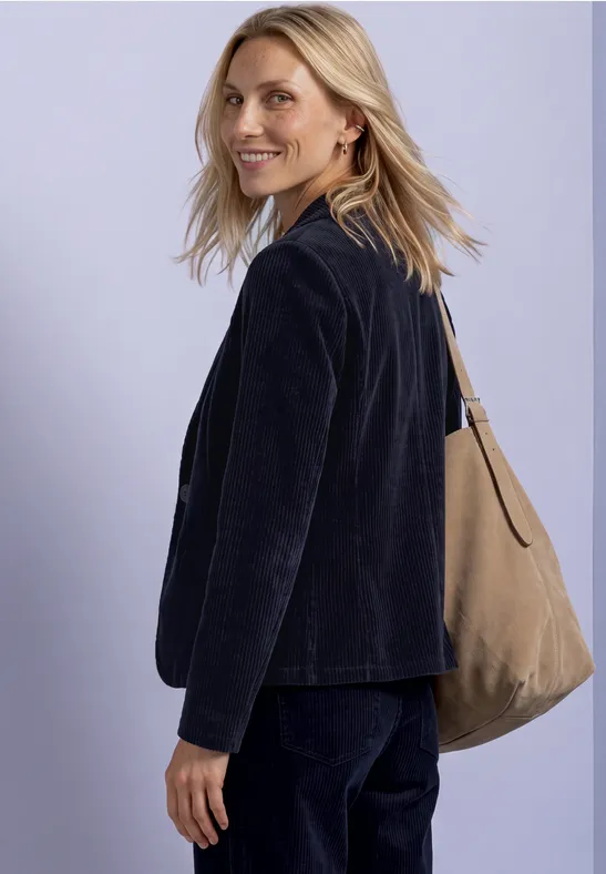 Een lachende blonde vrouw in een donkerblauwe outfit met een beige schoudertas voor een lichte achtergrond.
