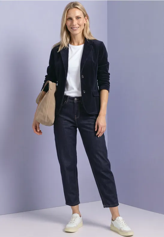 Vrouw in donkerblauw jasje, wit T-shirt, spijkerbroek en witte sneakers, met beige tas tegen lichtpaarse achtergrond.