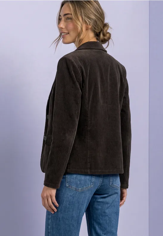 Een vrouw in jeans en een bruine corduroy jas kijkt glimlachend over haar schouder.