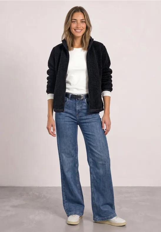 Eine Frau trägt eine schwarze Teddyjacke, weißen Pullover, weite Jeans und weiße Sneakers vor hellgrauem Hintergrund.