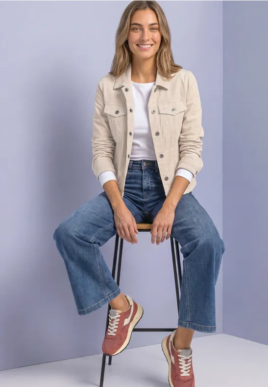 Eine Frau sitzt auf Hocker in Jeans, weißem Shirt, beiger Jacke und roten Sneakern vor violettem Hintergrund.