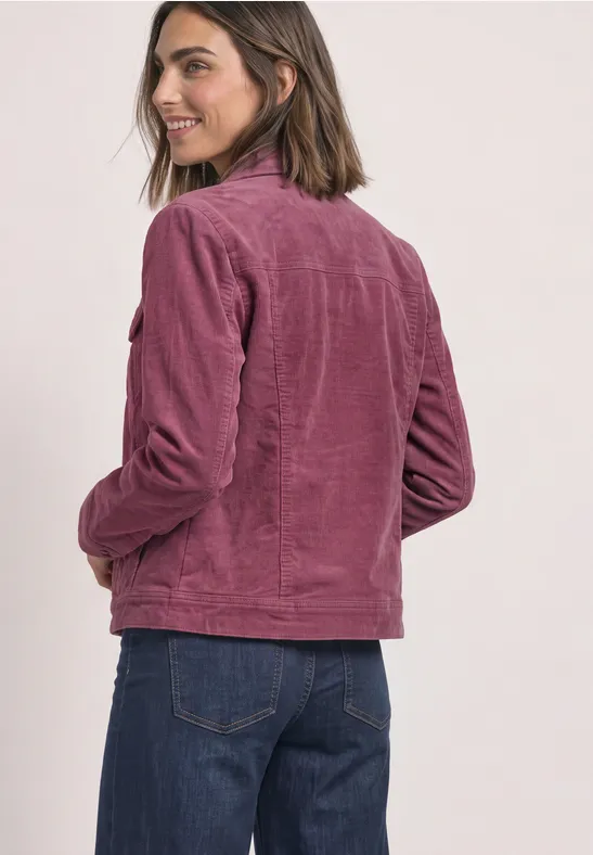 Junge Frau mit schulterlangen Haaren, trägt rosafarbene Cordjacke und blaue Jeans, lächelnd seitlich zur Kamera gewandt.