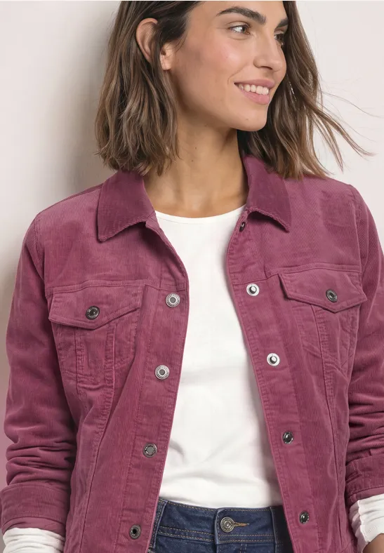 Frau in pinker Cordjacke mit weißem Shirt und Jeans lächelt seitlich.