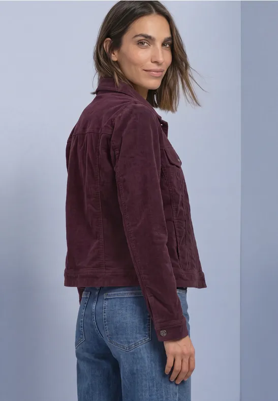 Frau in weinroter Cordjacke und blauen Jeans, steht seitlich und blickt zur Kamera vor schlichtem Hintergrund.