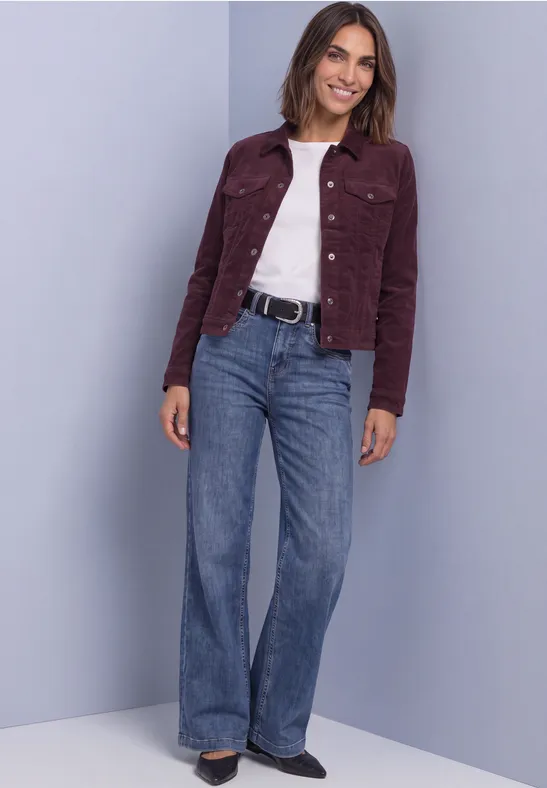 Frau in weinroter Cordjacke, weißem Shirt, weiten Jeans und Gürtel vor neutralem Hintergrund.