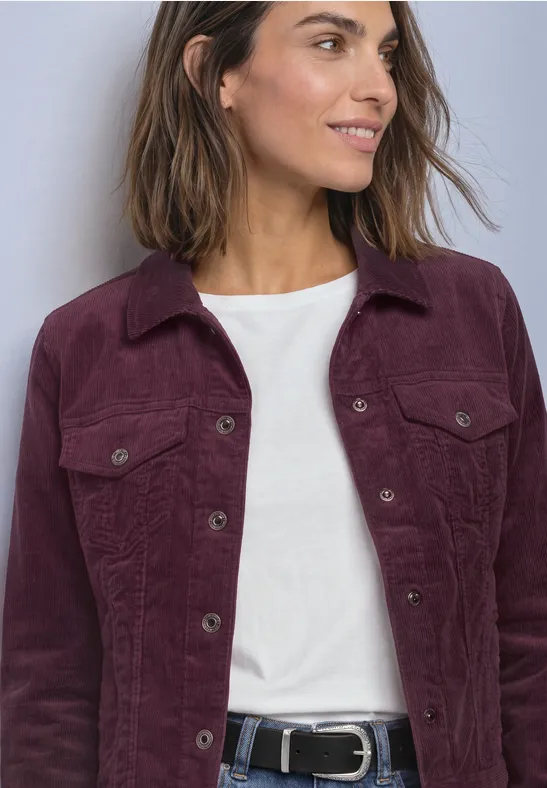 Frau trägt weiße Bluse unter lila Cordjacke mit Taschen und schwarzem Gürtel, lächelt seitlich.