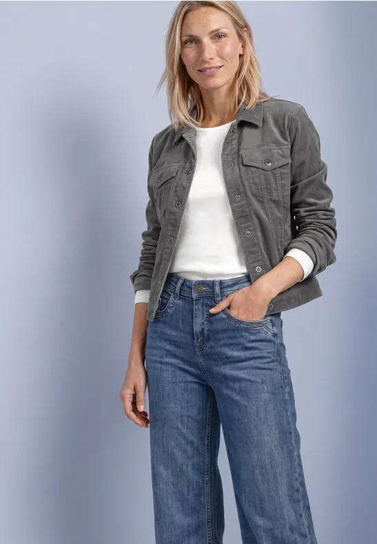 Femme en veste en velours côtelé vert foncé, haut blanc et jean bleu, souriante, main dans la poche.