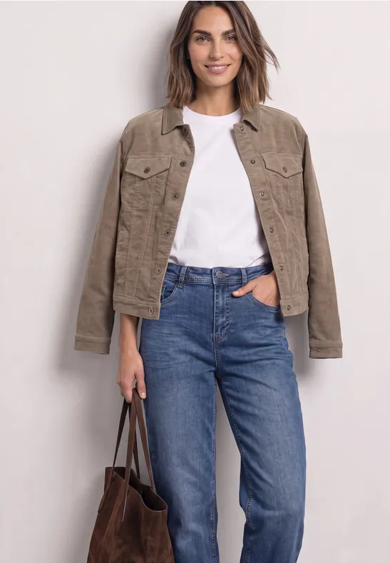 Frau trägt weißes T-Shirt, hellbraune Jacke, blaue Jeans und hält eine braune Tasche.