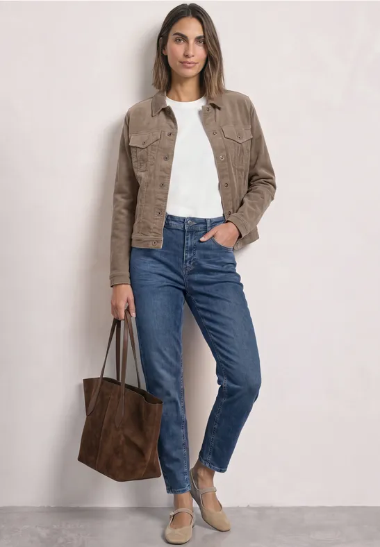 Frau in Jeans, weißem T-Shirt und brauner Jacke trägt eine große braune Tasche, steht vor neutralem Hintergrund.