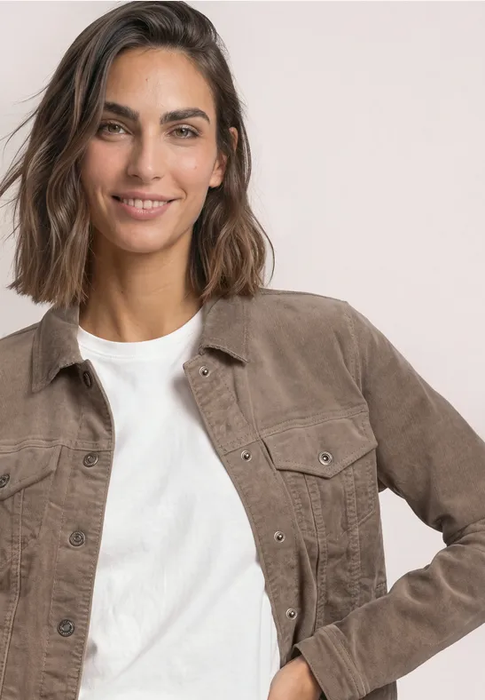Eine lächelnde Frau mit braunem, schulterlangem Haar trägt ein weißes Shirt und eine braune Jacke vor hellem Hintergrund.