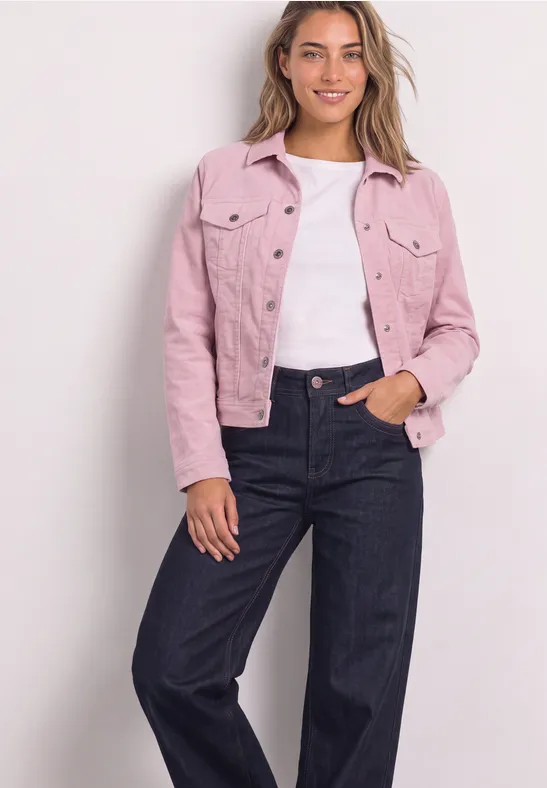 Une femme souriante porte une veste rose, un haut blanc et un jean foncé devant un mur clair.